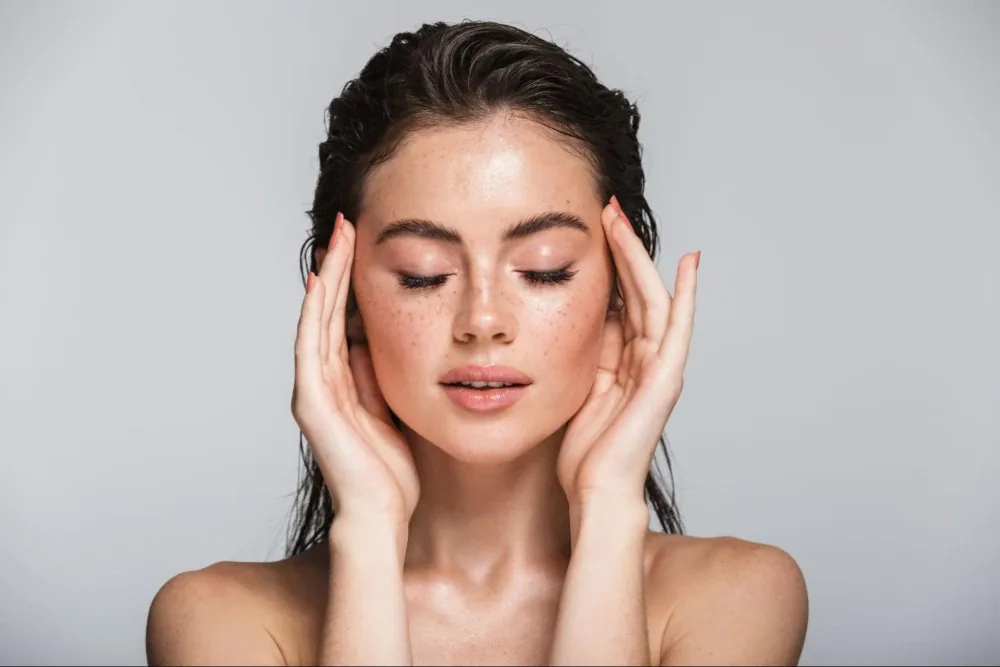 A woman with wet dark hair and freckles pressing her fingertips to her temples, eyes closed