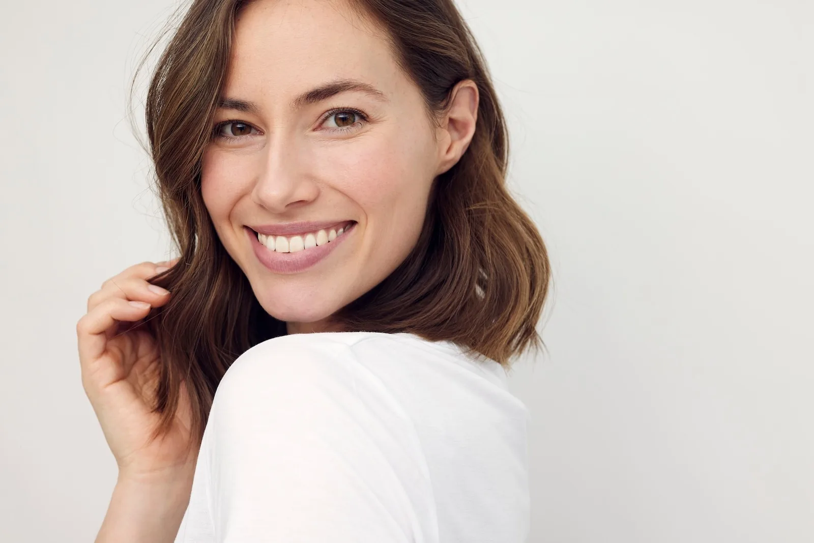 A smiling woman with shoulder-length brown hair in a white T-shirt looking over her shoulder