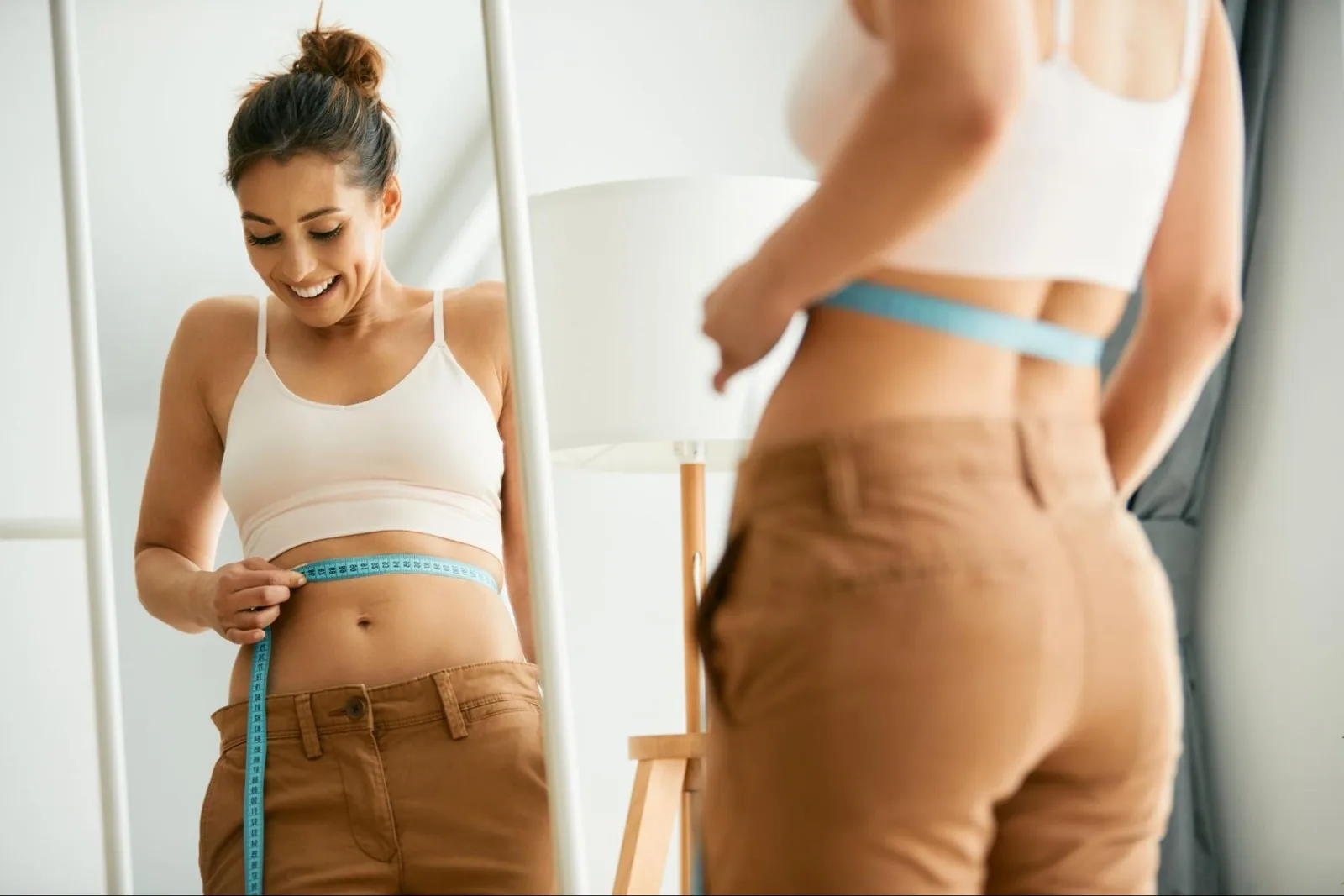 A smiling woman measuring her waist with a blue tape measure in front of a mirror