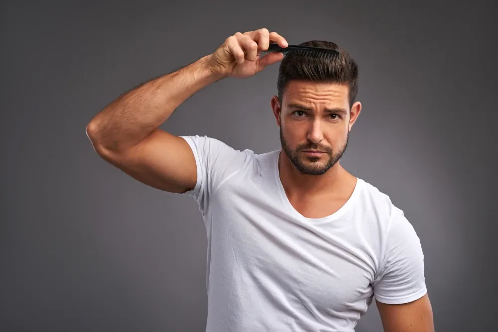 A bearded man in a white T-shirt combing his hair and frowning at the camera