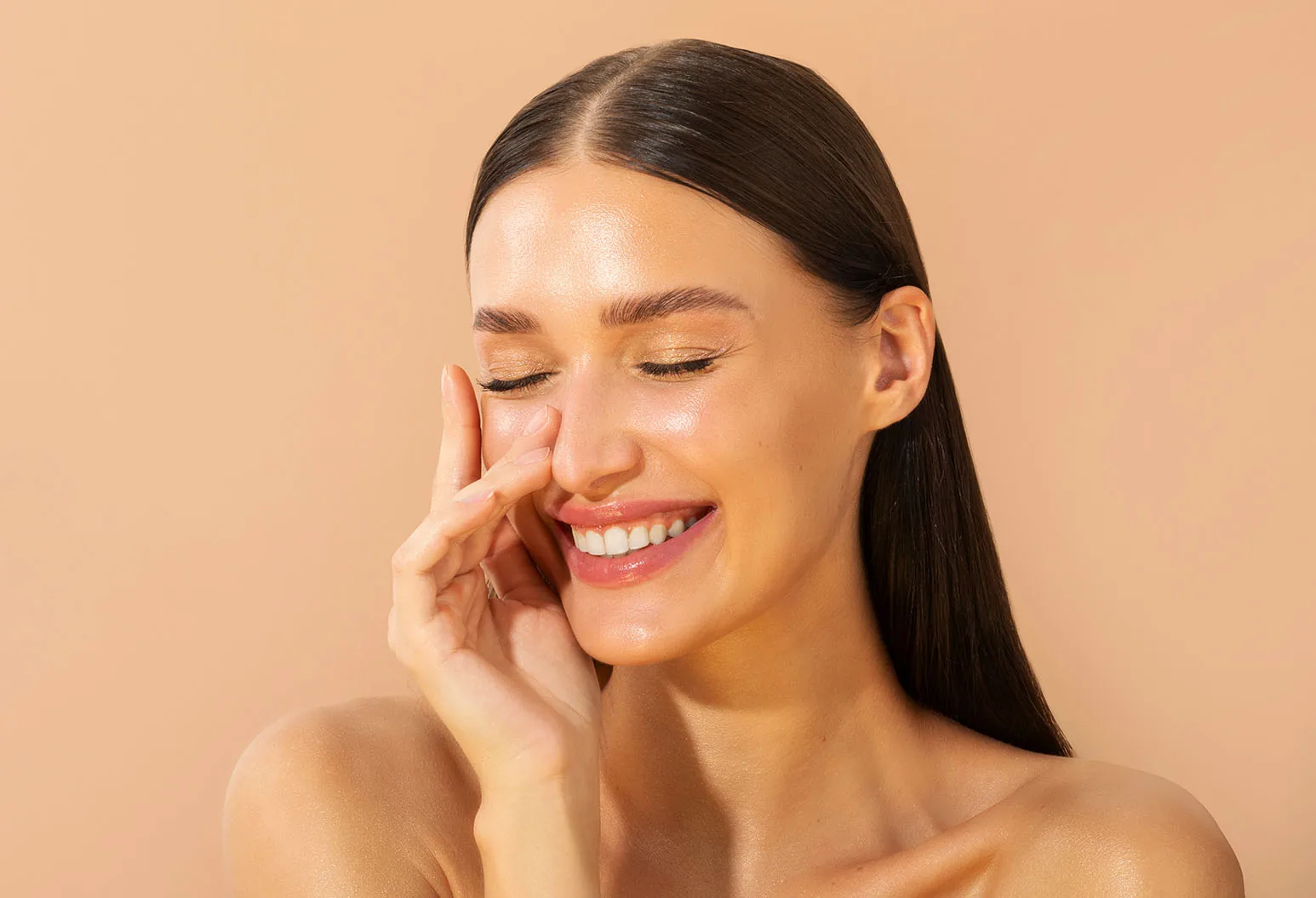 A laughing woman with dark hair and eyes closed touching her cheek against a peach background