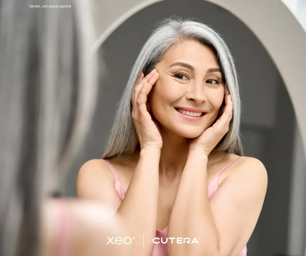 A woman with long grey hair smiling at her reflection while touching her cheeks