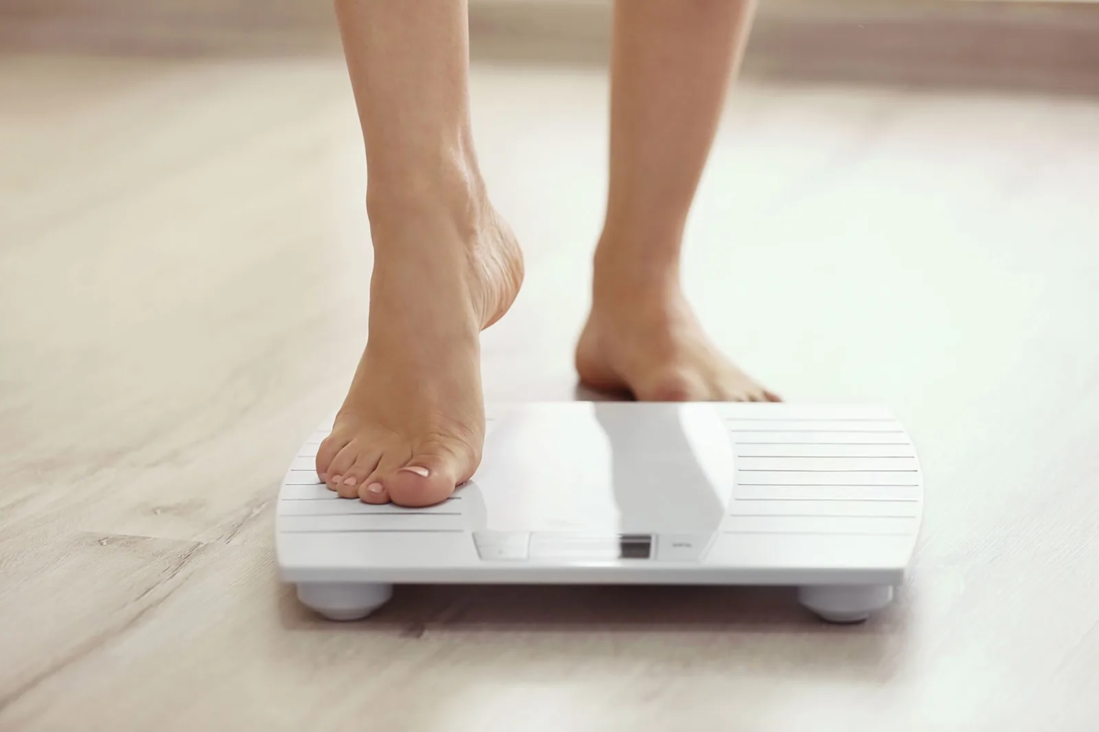 Bare feet stepping onto a white digital bathroom scale on a light wooden floor