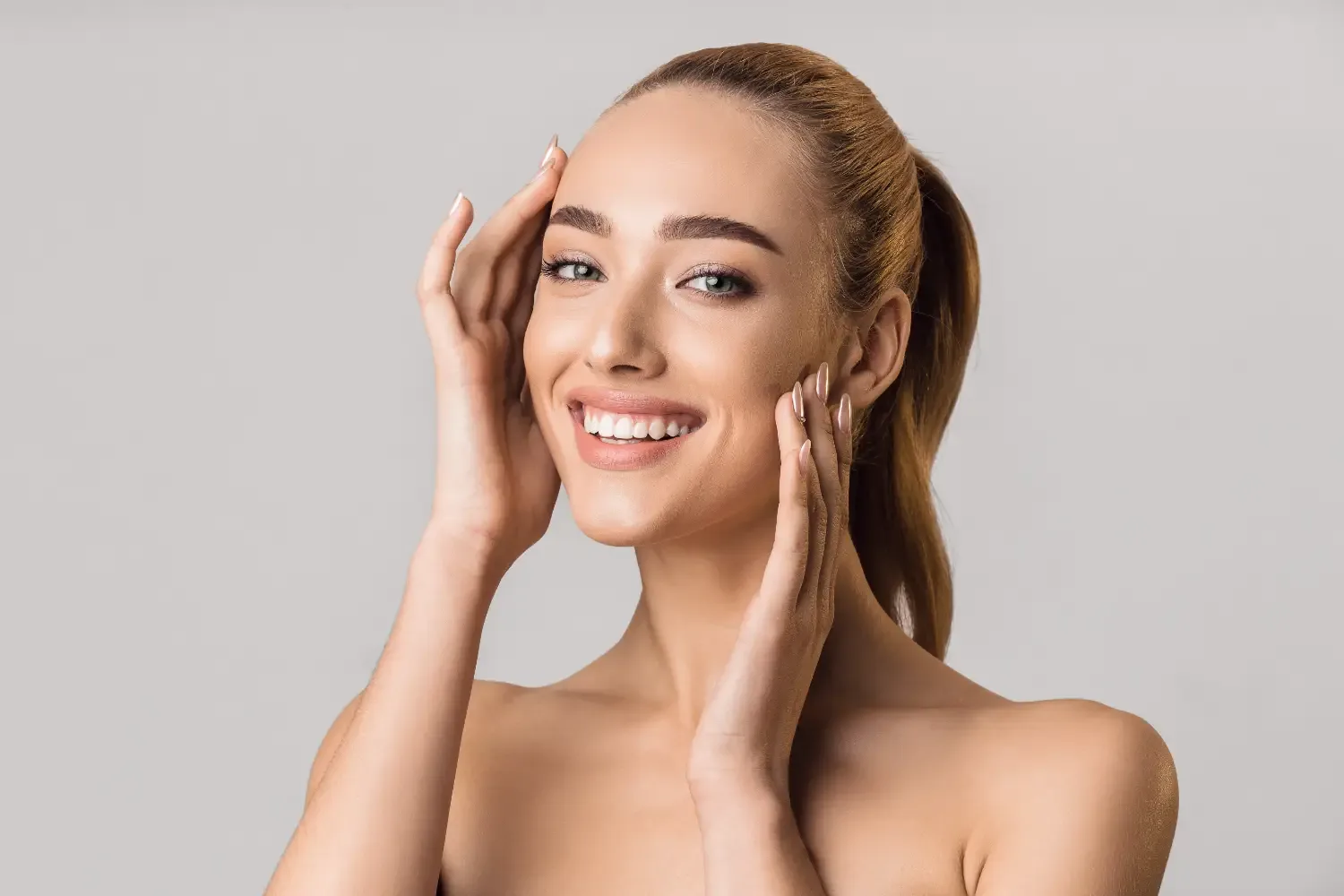 A smiling woman with a high ponytail holding her fingertips to her temple and cheek