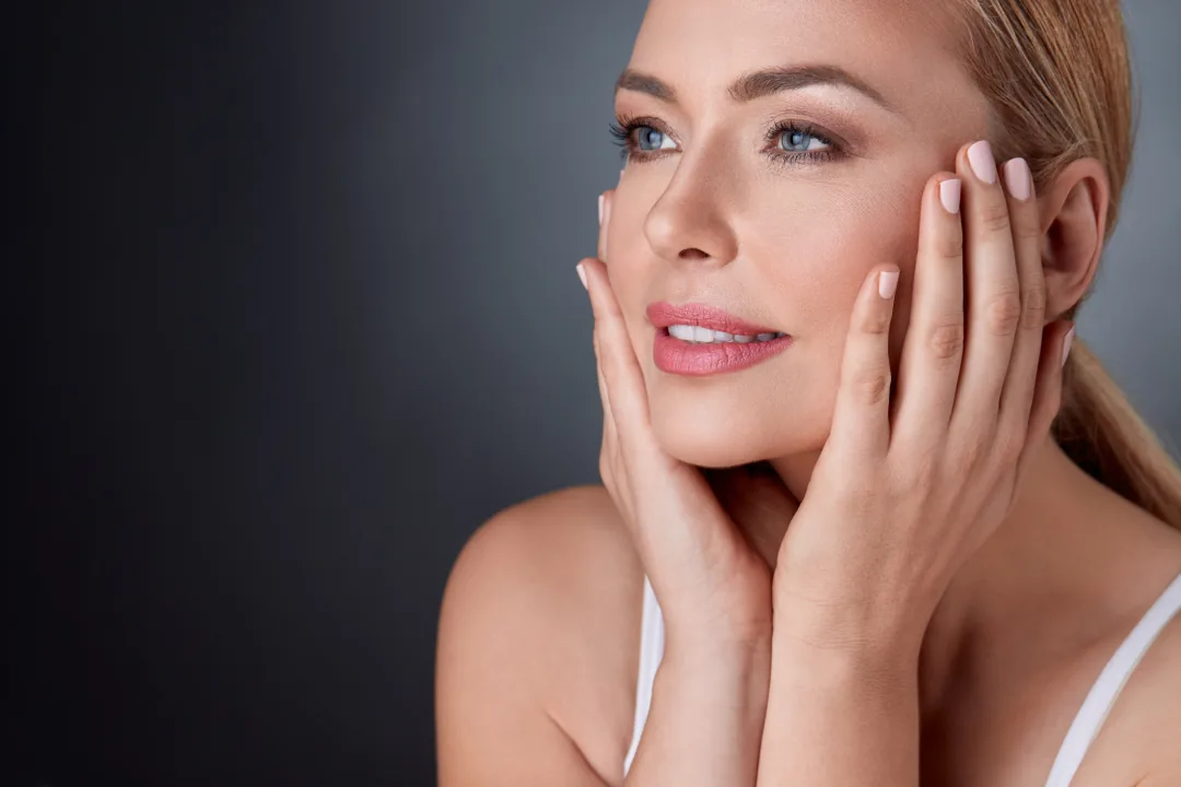 A blonde woman cupping her face in both hands and gazing upward against dark grey