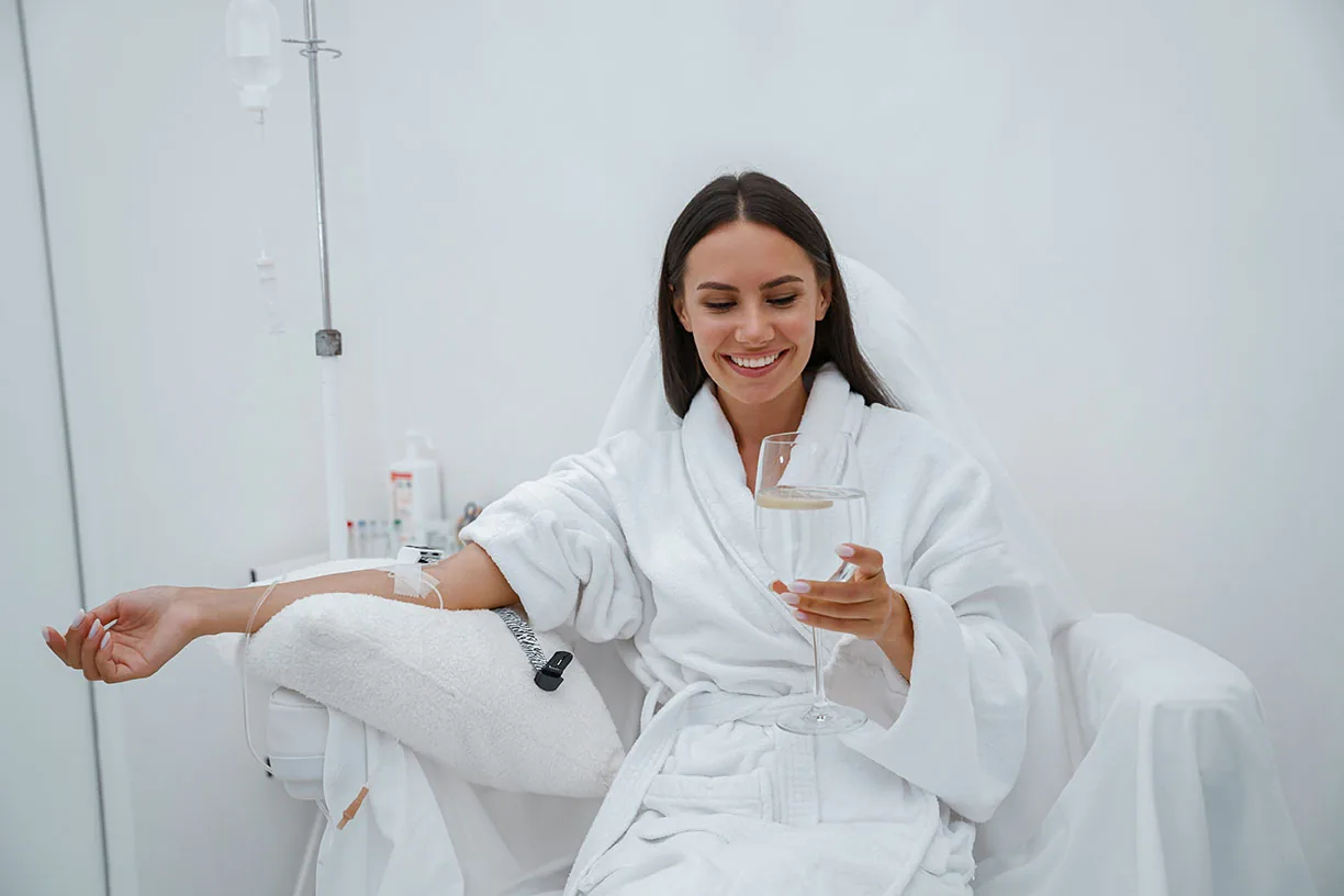 A smiling woman in a white bathrobe holding a glass while receiving an IV drip