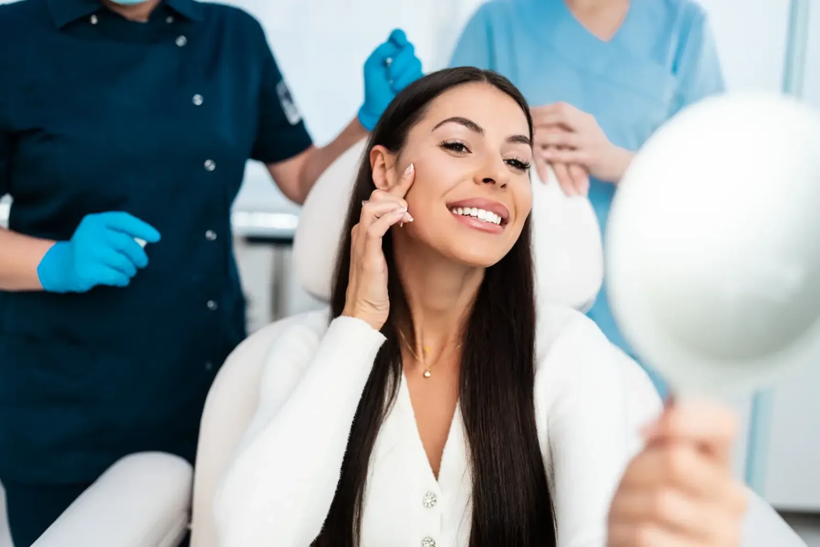 A smiling woman holding a hand mirror to her face with two gloved staff behind her