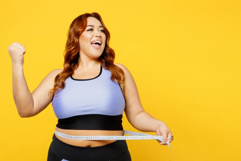 A red-haired woman in a lilac sports top cheering with a tape measure at her waist
