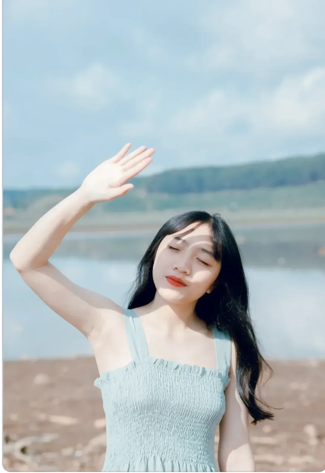 A young woman with long dark hair shielding her eyes from the sun beside a lake