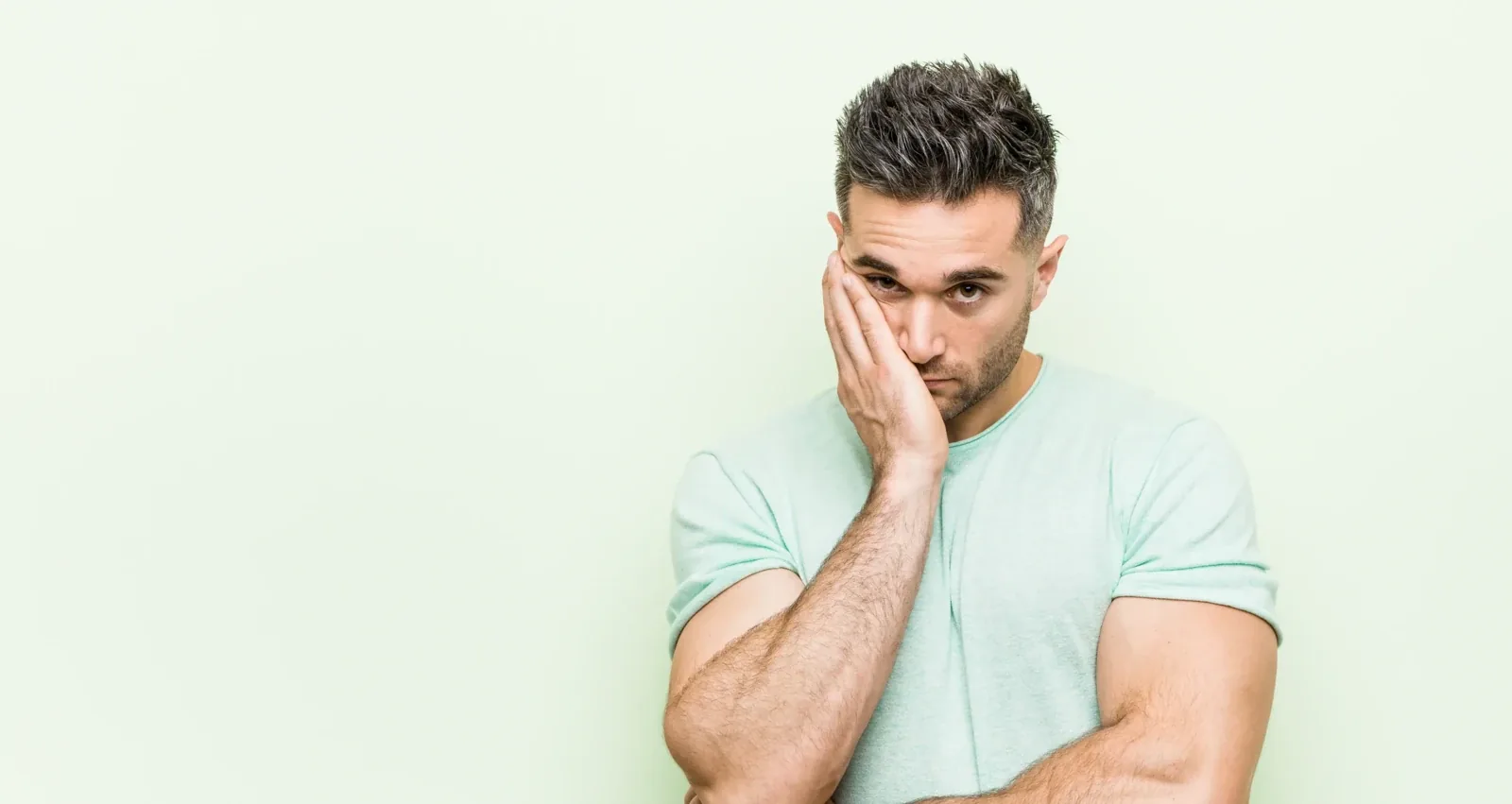 A weary man in a mint T-shirt resting his cheek on his hand, arms crossed