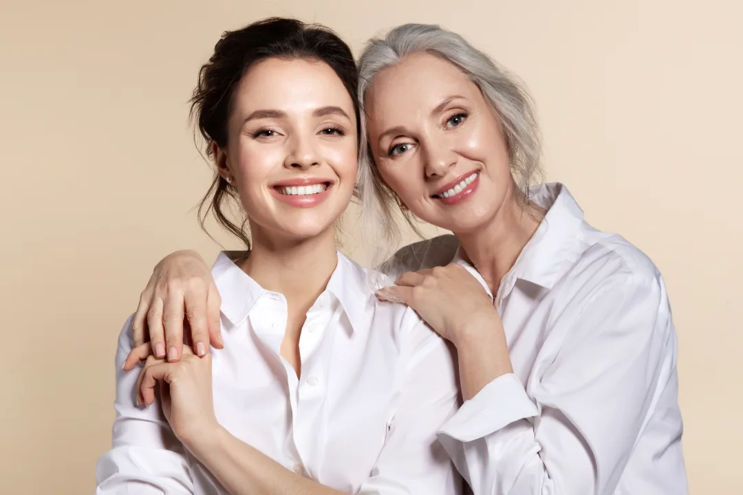Two smiling women in white shirts, one young and one grey-haired, embracing