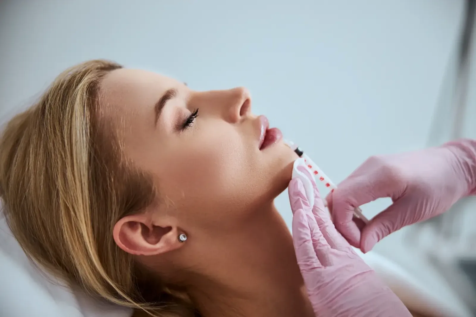 Pink-gloved hands holding a syringe to the chin of a reclining blonde woman with eyes closed