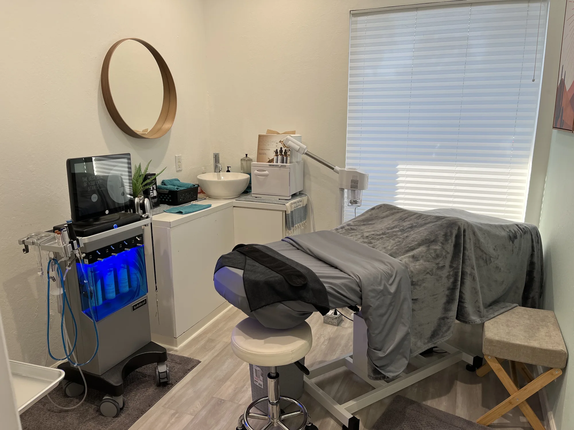 The facial room at Radiance Wellness &amp; Aesthetics, with a round mirror, a treatment bed and the HydraFacial machine