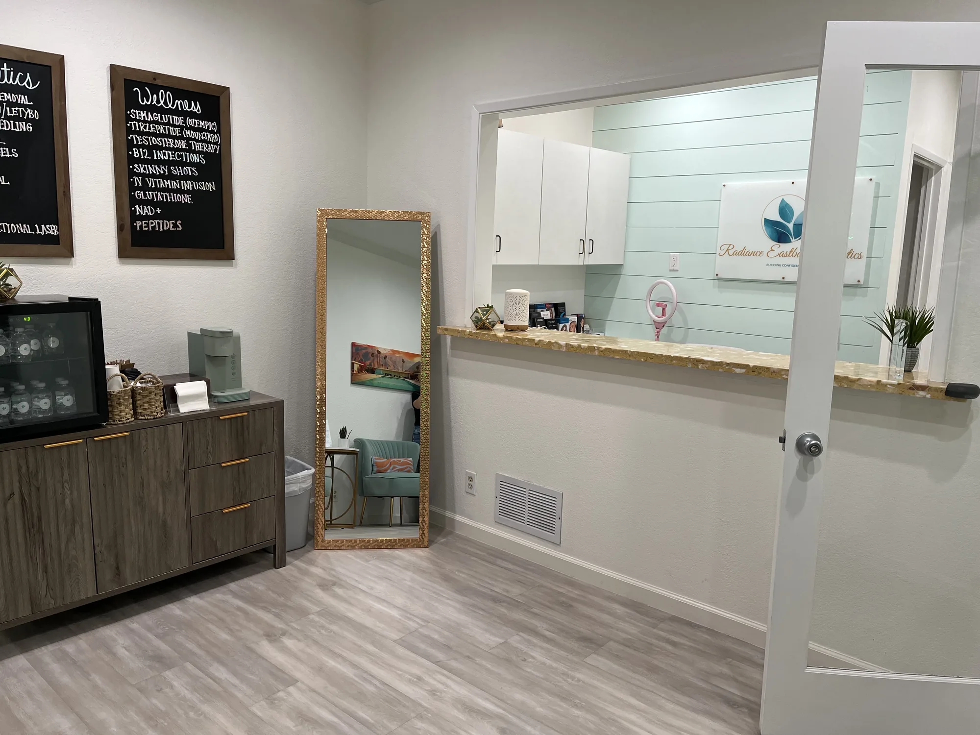 The front desk at Radiance Wellness &amp; Aesthetics with the aesthetics and wellness menu boards and a gold-framed mirror