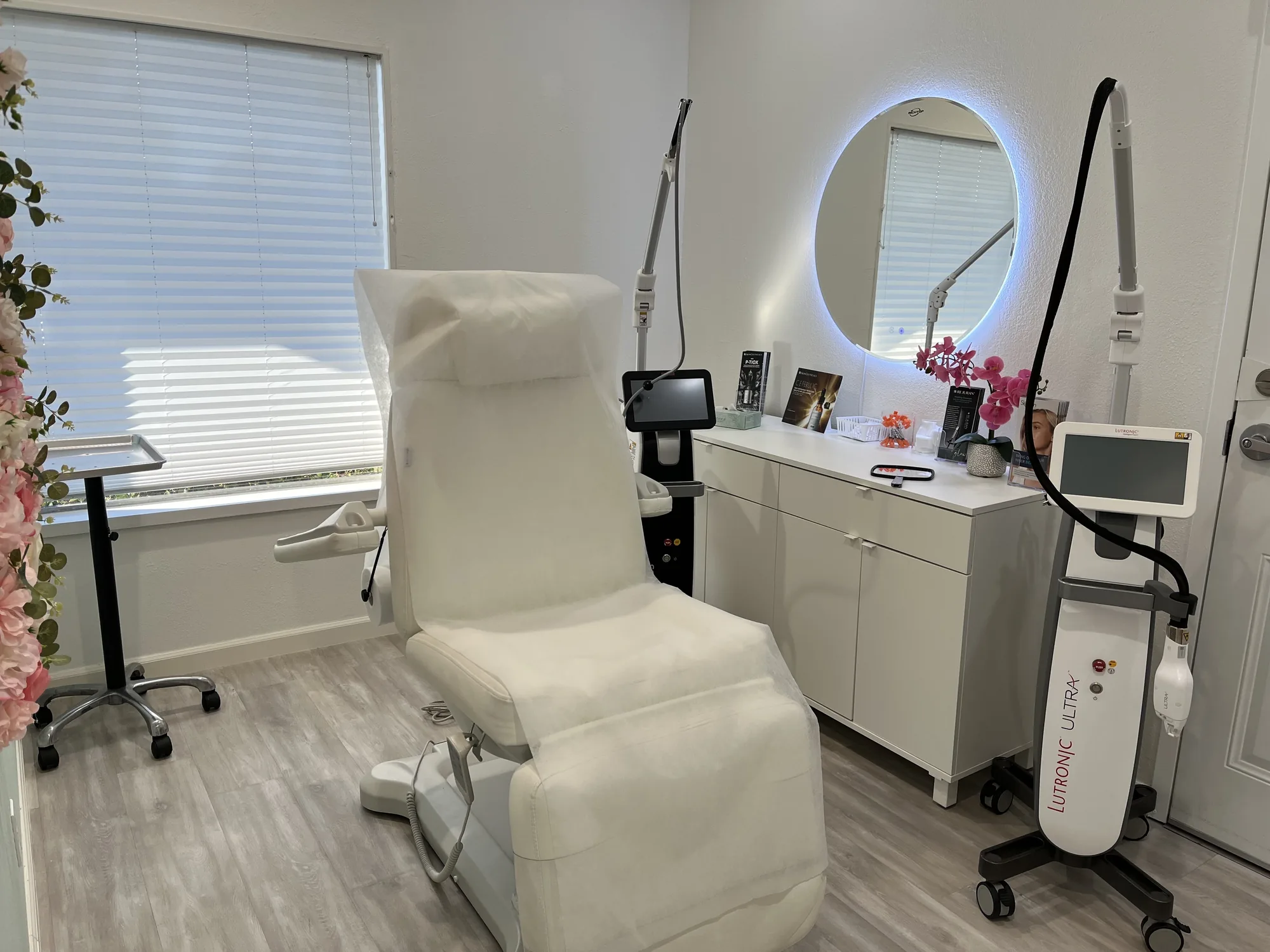 A treatment room at Radiance Wellness &amp; Aesthetics with a white treatment chair, a ring-lit mirror and a laser device